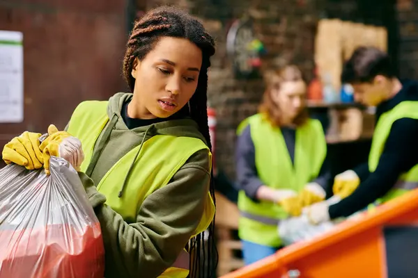 A young woman in a green vest and gloves holding a bag of garbage, participating in a trash sorting activity with other eco-conscious volunteers. — Stock Photo