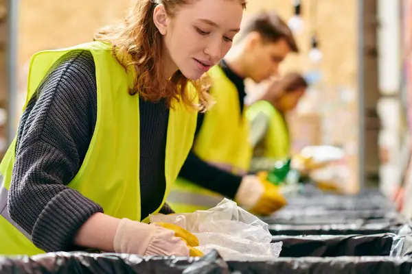A young woman in a yellow safety vest sorts trash at a table with other eco-conscious volunteers. — Fotografia de Stock
