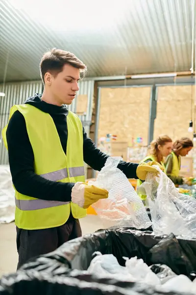 A young man in a yellow safety vest diligently picks up trash with other eco-conscious volunteers in gloves. — Stock Photo