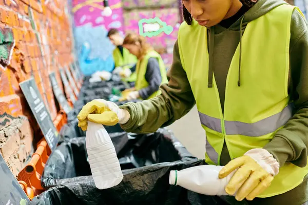 Young volunteer in green jacket and yellow gloves sorts trash with eco-conscious people wearing safety vests. — Stock Photo