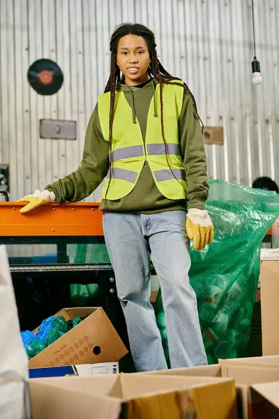 A young african american volunteer in gloves and a safety vest sorts waste in a warehouse, demonstrating eco-conscious behavior. — Stock Photo