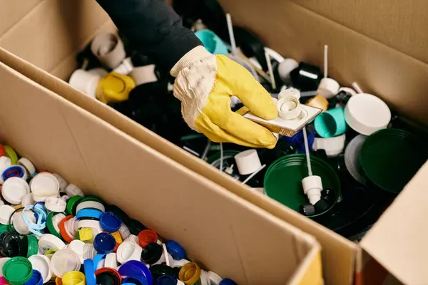 A box overflows with cups of various colors, sorted by a young volunteer in gloves and safety vest for recycling. - foto de stock