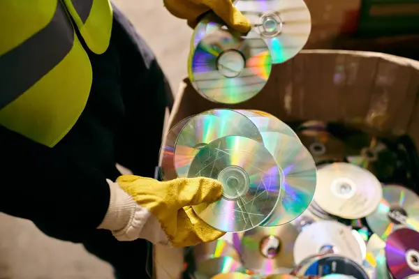 A young volunteer in gloves and safety vest holding a bunch of CDs, sorting waste as part of eco-conscious efforts. — стоковое фото