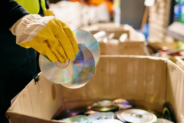 A person in yellow gloves carefully holds a CD inside a box, sorting waste for a cleaner environment. — Foto stock