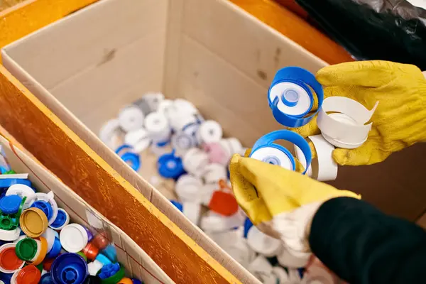 A young volunteer in a yellow glove carefully holds a pair of matching gloves over a box of waste, showing eco-consciousness. — Stock Photo
