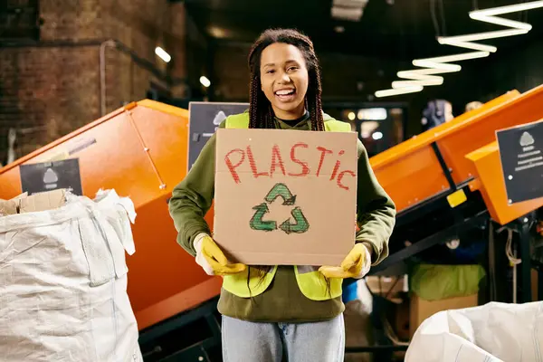 Young volunteer in safety gear sorting waste, holding a sign that says plastic. - foto de stock