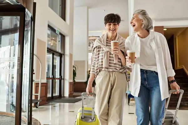 Due donne lesbiche anziane camminano felicemente lungo un corridoio di un hotel con i loro bagagli. — Foto stock