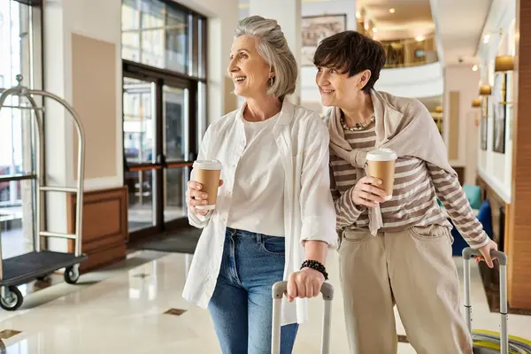 Senior lesbiana pareja camina por hotel pasillo con equipaje. - foto de stock