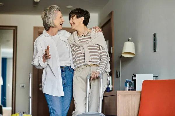 Two women, a loving senior lesbian couple, stand in a cozy living room with their luggage. — Stock Photo
