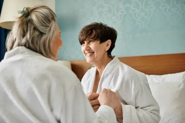 Una mujer con bata de baño sonríe mientras otra mujer se pone su bata en un tierno momento de conexión. - foto de stock