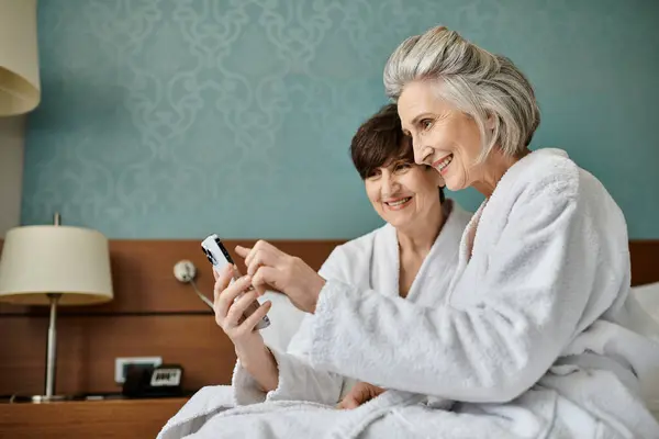 Старшая лесбийская пара в любящих объятиях на кровати, поглощенная экраном мобильного телефона. — стоковое фото