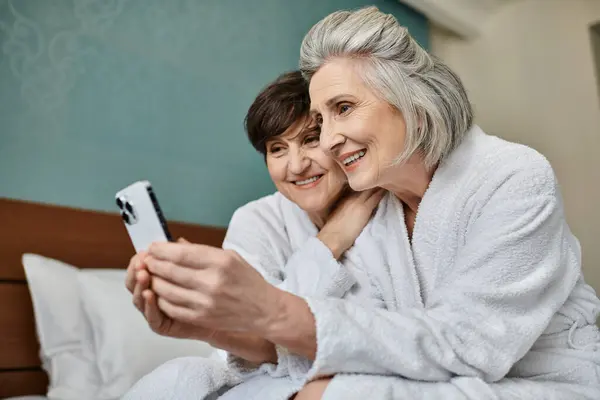 Senior lesbian couple share a tender moment while viewing a cell phone on a bed. — Stock Photo