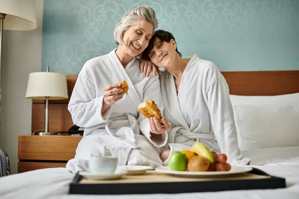 A senior lesbian couple in robes, seated on a bed, exuding tenderness and love. — Stock Photo