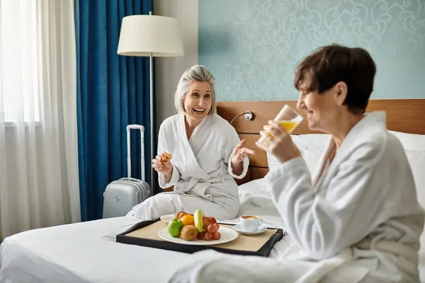 Two elderly women share a tender moment while sitting on top of a cozy hotel bed. — Stock Photo