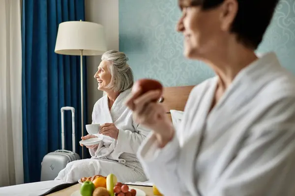 Two senior women in a loving embrace, sitting together on top of a bed in a hotel room. — Stock Photo