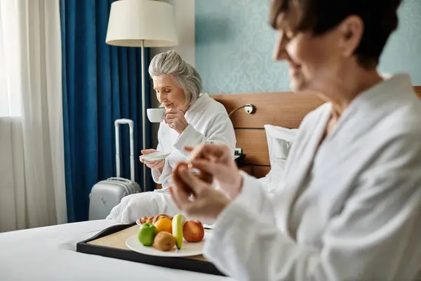 Dos mujeres lesbianas mayores con túnicas blancas, tiernamente sentadas en una cama. - foto de stock