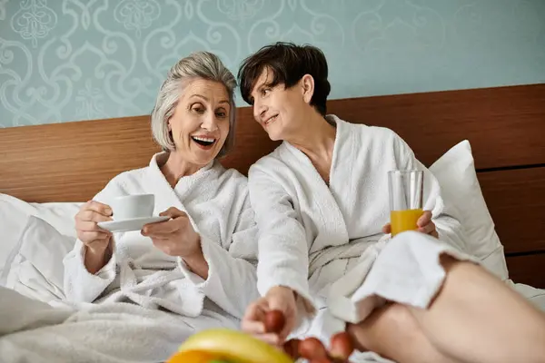 Two women in white robes, a senior lesbian couple, sit peacefully on a bed. — Stock Photo