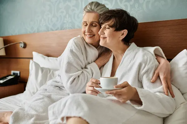 Due lesbiche anziane in accappatoio condividono un momento tenero su un letto, uno in possesso di una tazza. — Foto stock