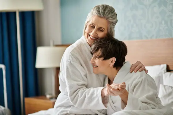A senior lesbian couple sharing a tender moment in a bed, with a woman in a white robe embracing her partner. — Stock Photo
