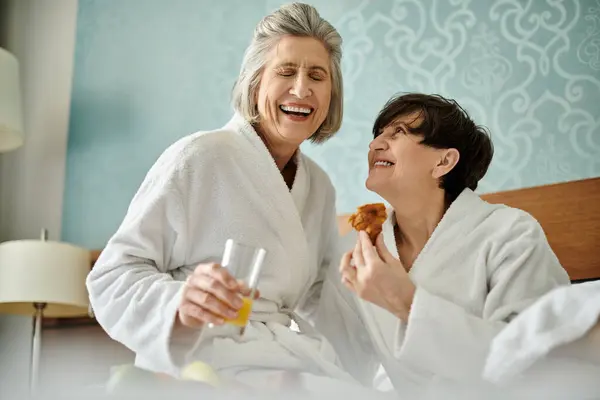 Dos mujeres lesbianas senior con túnicas blancas comparten una comida junto con amor y ternura. - foto de stock
