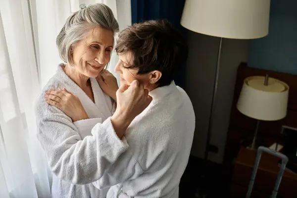 Una pareja de lesbianas mayores en albornoces compartiendo un tierno abrazo en un hotel. - foto de stock
