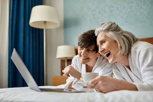 Two loving lesbian partners, one older and one younger, sharing a tender moment on a bed while using a laptop. — Stock Photo