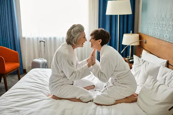 Senior lesbian couple peacefully sitting together on top of a bed in a hotel room. — Stock Photo