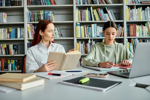 A redhead tutor and a teenage girl, sit at a library table. The tutor teaches while using a laptop for modern education. — Stock Photo