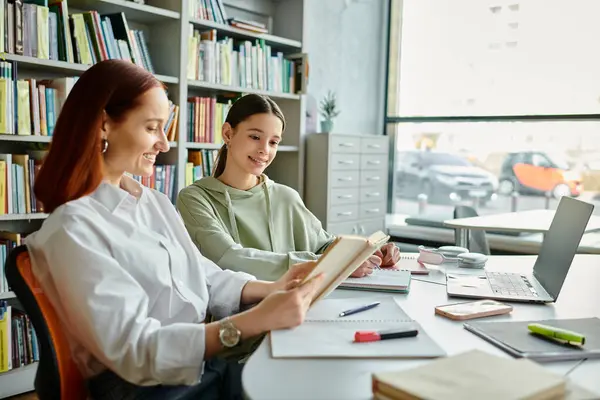A redhead tutor is teaching a teenage girl in a library, having after-school lessons while using a laptop. — Stock Photo