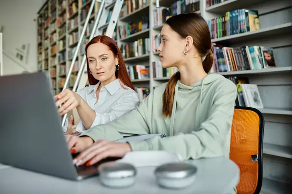 A tutor with red hair guides a student at a library, both working on a laptop for after-school lessons. Modern education in action. — Stock Photo