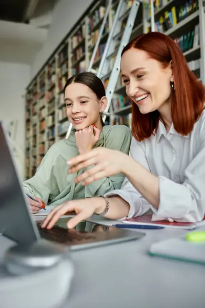 Un tutor rossa insegna una ragazza adolescente in una biblioteca, sia intensamente lavorando su un computer portatile per la moderna educazione post-scolastica. — Foto stock