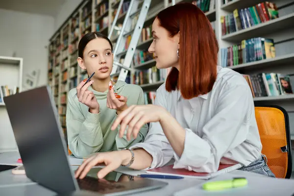 Un tutor rossa insegna una ragazza adolescente in una biblioteca, entrambi assorbiti in un computer portatile durante una lezione dopo la scuola. — Foto stock