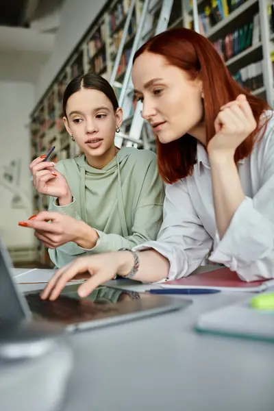 A tutor with red hair guides a teenage student in a library, both engrossed in the laptop screen for after-school lessons. — Stock Photo