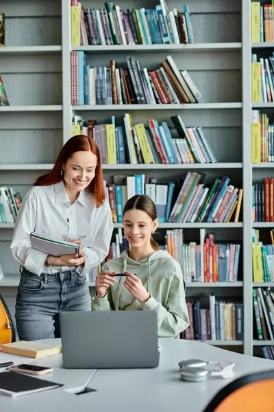 Рыжеволосый репетитор преподает девочке-подростку в библиотеке, обе занимаются ноутбуком во время занятий после школы.. — стоковое фото