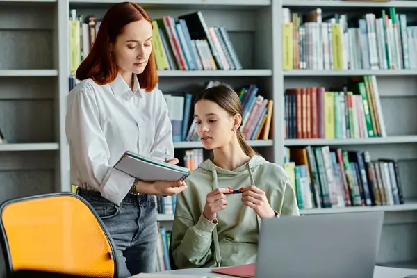 A redhead tutor is teaching a teenage girl in a library, with a laptop in front of them for their modern after-school lessons. — Stock Photo