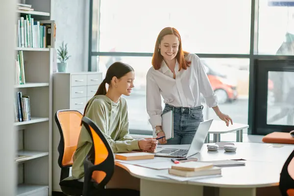 Un tutor rossa guida uno studente adolescente attraverso lezioni post-scolastiche presso una scrivania in un ufficio moderno, entrambi immersi nello schermo del computer portatile. — Foto stock