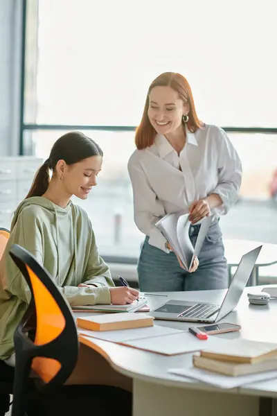 A redhead tutor guides a teenage girl through after-school lessons at a desk, both using laptops for modern education. — Stock Photo