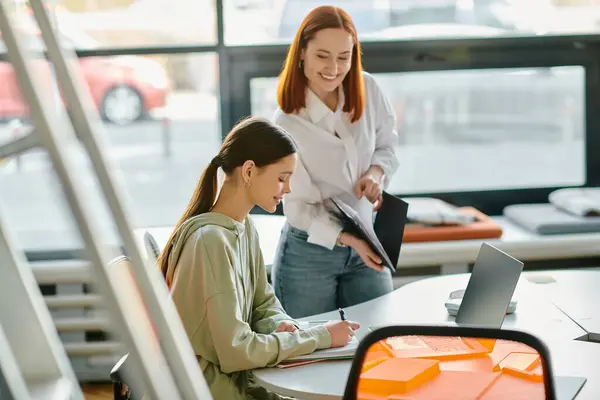 A redhead tutor leads after-school lessons with a teenage girl at a table in an office, utilizing a laptop for modern education. — Stock Photo