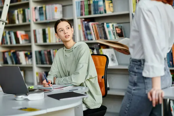 Un tutor dai capelli rossi insegna a un'adolescente in una biblioteca, entrambe sedute a una scrivania, utilizzando un computer portatile per l'educazione moderna.. — Foto stock