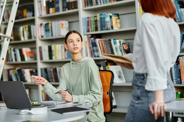 Un tutor pelirrojo enseña a una adolescente en una biblioteca, ya que participan en lecciones después de la escuela usando una computadora portátil. - foto de stock