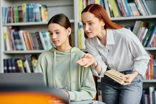 Redhead tutor helping a teenage girl in a library, both engrossed in learning on a laptop. — Stock Photo