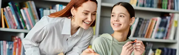 A redhead tutor engages in discussions with a teenage girl in a bustling library setting, using a laptop for modern education. — Stock Photo