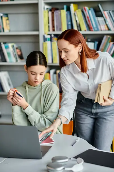 A redheaded tutor teaches a teenage student in a library, both focused on the laptop screen. — Stock Photo
