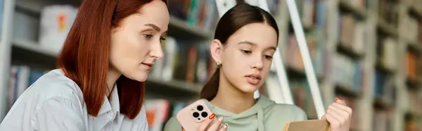 Un tutor rossa e una ragazza adolescente, assorto nei loro telefoni in una biblioteca. — Foto stock