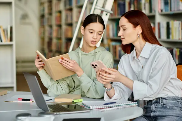 A young tutor with red hair teaches a teenage girl at a library table, utilizing a laptop for after-school lessons. — Stock Photo