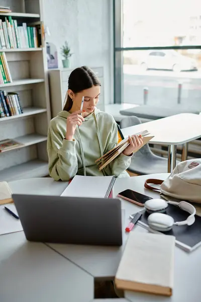 Una adolescente profunda en el pensamiento, multitarea entre su portátil y un libro en el escritorio mientras hace la tarea después de la escuela. - foto de stock