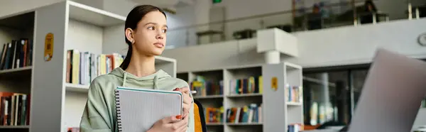 Девочка-подросток стоит перед книжным шкафом в библиотеке, погруженная в учебу и домашнее задание на ноутбуке после школы. — стоковое фото