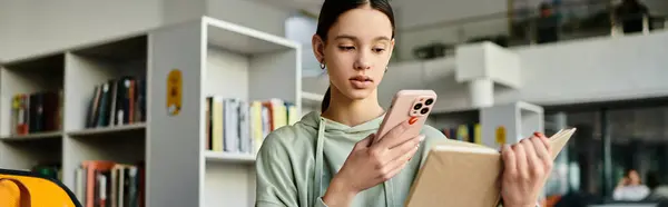 Ein Mädchen im Teenageralter hält ein Buch in der Hand, während sie ihr Telefon checkt, vertieft in moderne Bildung und balanciert ihr Studium mit Technologie. — Stockfoto