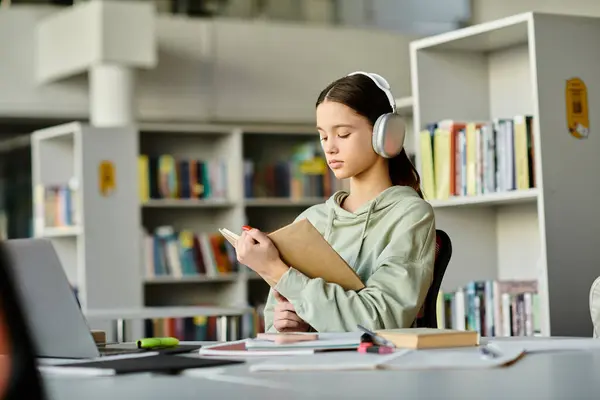 Un'adolescente con le cuffie si siede ad una scrivania in una biblioteca, immersa nel fare i compiti sul suo portatile dopo la scuola. — Foto stock