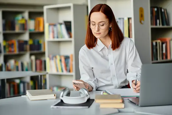 Una tutor rossa conduce lezioni online, seduta a una scrivania in una biblioteca con un computer portatile. — Foto stock